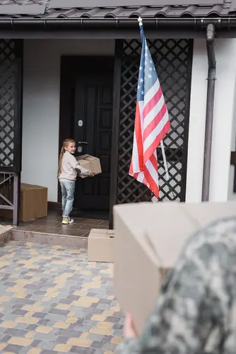 Young girl carrying a cardboard box to the front door of a house, with an American flag displayed outside and a person in military camouflage holding another box in the foreground.