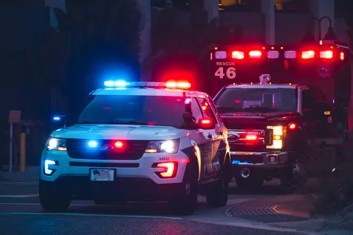A police SUV with flashing red and blue lights is parked in front of a rescue ambulance vehicle at night. Both emergency vehicles are illuminated and appear to be responding to an incident.