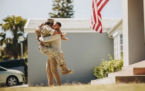 U.S. military service member in uniform joyfully embracing and being lifted off the ground by a loved one outside a home with an American flag hanging nearby.