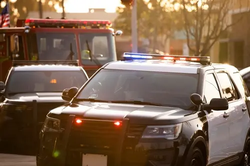 Police SUV with red and blue lights flashing, parked near a fire truck and another police vehicle in an urban setting at sunset.