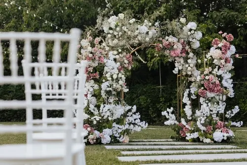 Outdoor wedding ceremony setup featuring a floral arch adorned with white, blush, and pink flowers, with white chairs arranged in rows on a grassy lawn.
