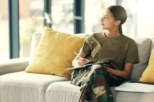 Female service member in military fatigues sitting on a sofa with a notebook and pen, looking thoughtfully out a window.