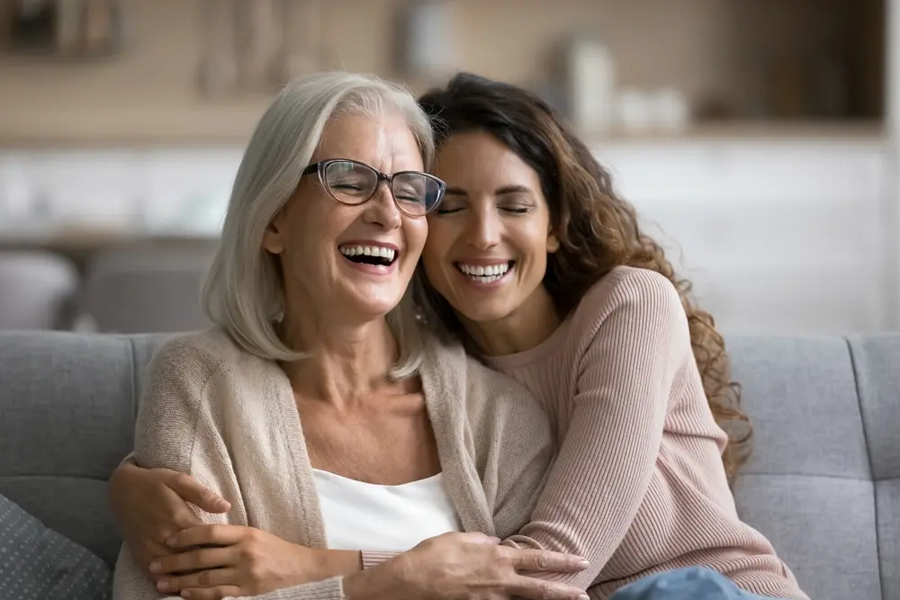 A joyful older woman with gray hair and glasses sits on a couch, laughing and hugging a younger woman with long curly hair. Both are smiling with eyes closed, sharing a warm moment in a cozy indoor setting.