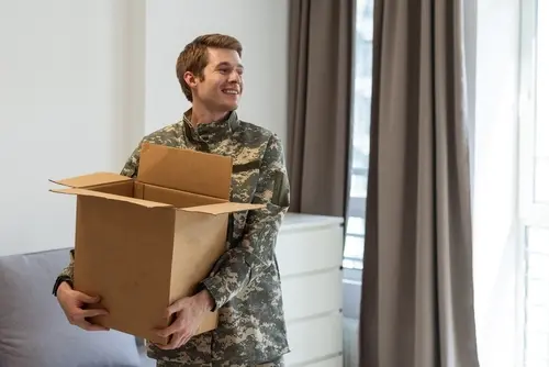Smiling male U.S. service member in camouflage uniform holds an open cardboard box inside a bright room, possibly in the process of moving in or out.